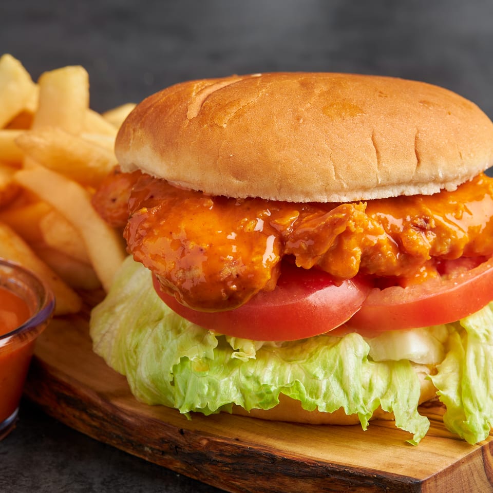 Spicy fried chicken sandwich with lettuce and tomato, served with fries and dipping sauce on a wooden board.