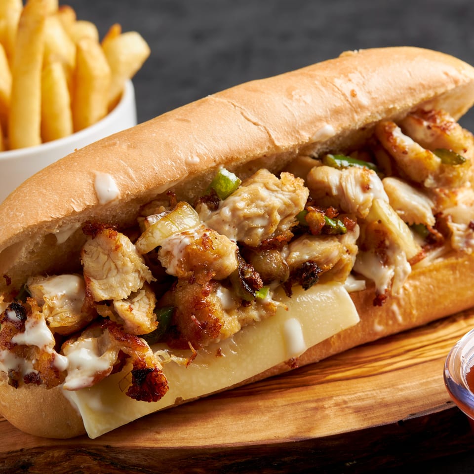 Grilled chicken sandwich with melted cheese, peppers, and creamy sauce, served with fries and dipping sauce on a wooden board.