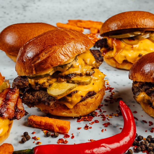 Multiple Hip Hop Fish & Chicken cheeseburgers with pickles, bacon, and chili flakes on a white surface.