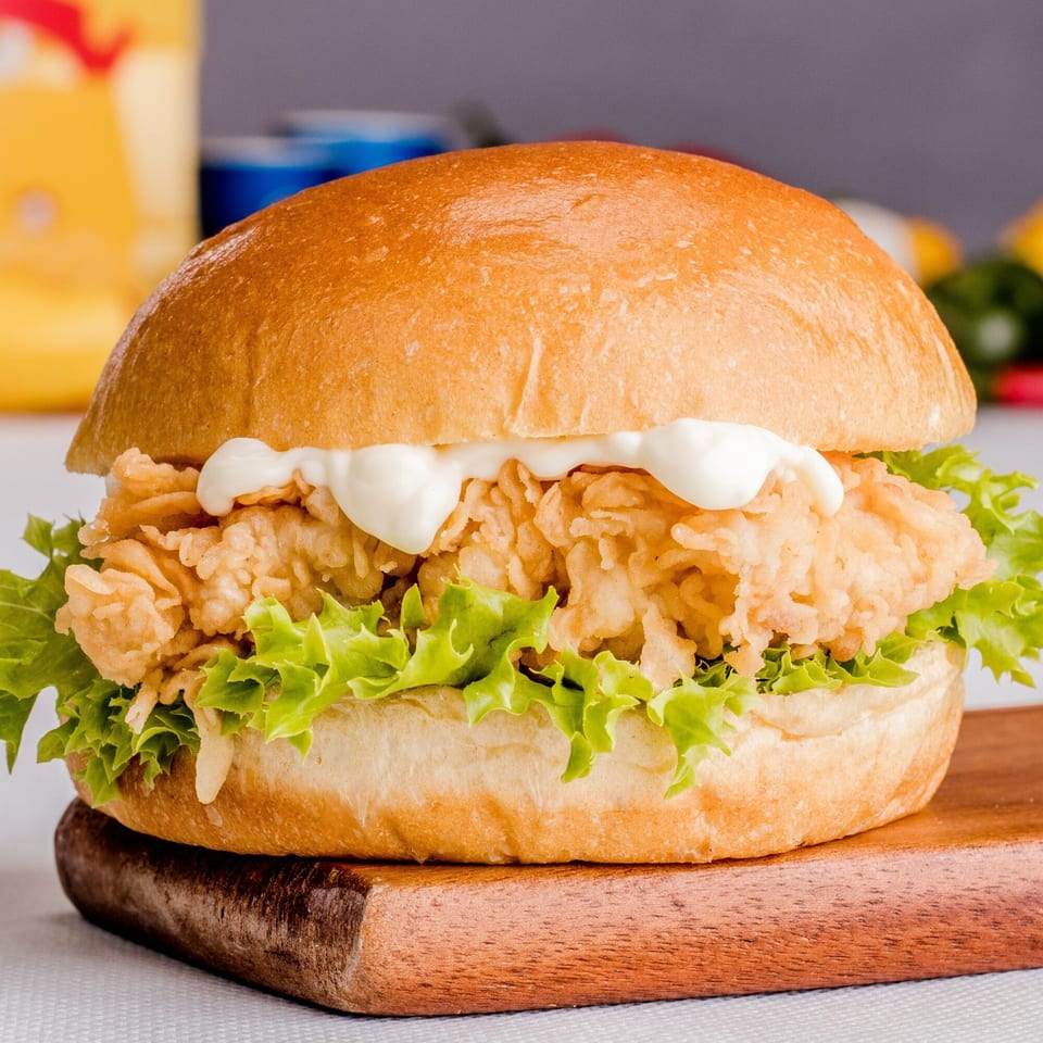 Crispy fried chicken sandwich with lettuce and mayonnaise on a bun, served on a wooden board.