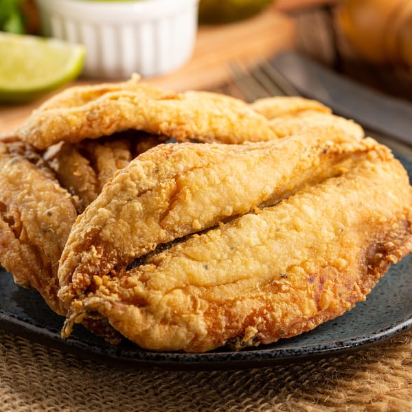 Crispy golden-brown fried fish fillets on a dark plate, with a lime wedge and sauce in the background.