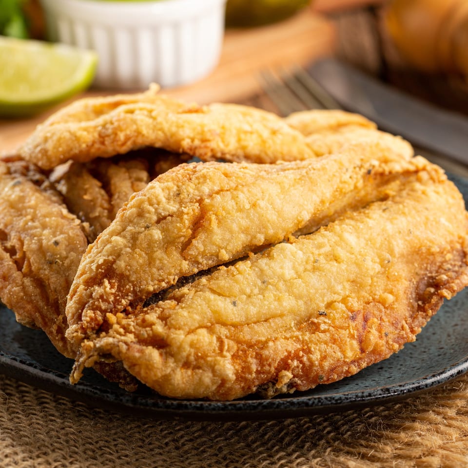 Crispy golden-brown fried fish fillets on a dark plate, with a lime wedge and sauce in the background.
